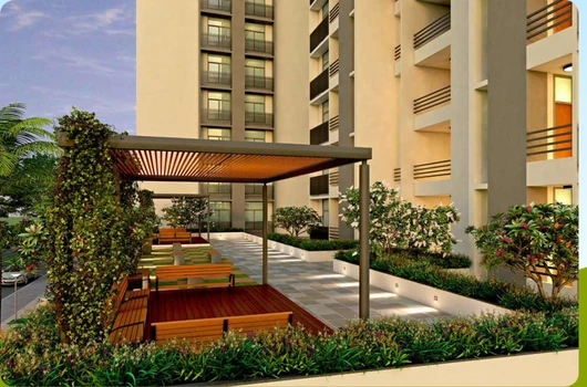 A beautiful outdoor seating area with lush greenery, part of the amenities offered in affordable flats in ahmedabad under 30 lakhs by Laxmi Group. Features include 24/7 security, CCTV surveillance, children's play area, indoor games, and a multipurpose hall.